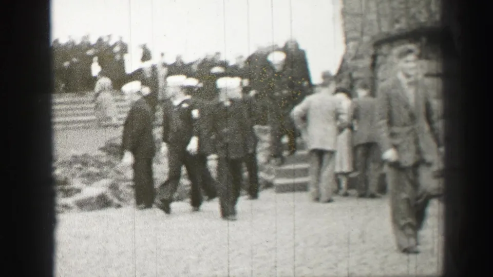 Grainy 1952 Scotland frame with uniformed men and civilians moving toward crowded outdoor stone steps.