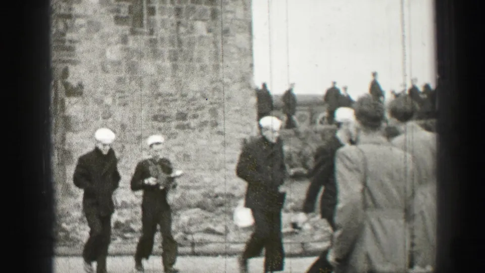 Uniformed sailors in white caps move near a rough stone wall in Edinburgh, with civilians and steps visible in the background.