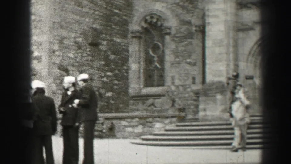 1952 Scotland home-movie frame showing suited men in hats gathered near stone cathedral steps and an ornate window.