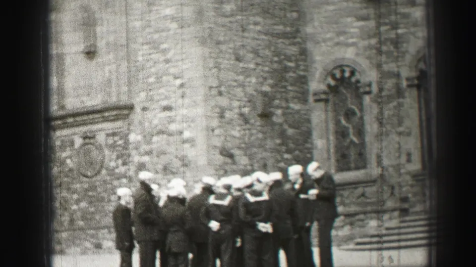 The handheld 8mm frame shows sailors in white caps gathered near a Gothic-style church wall in Scotland.