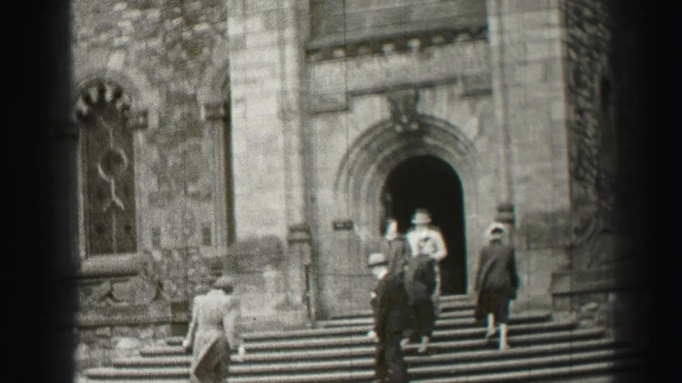 Black-and-white 8mm frame of adults in coats and hats approaching the arched entrance of a stone church in Edinburgh.