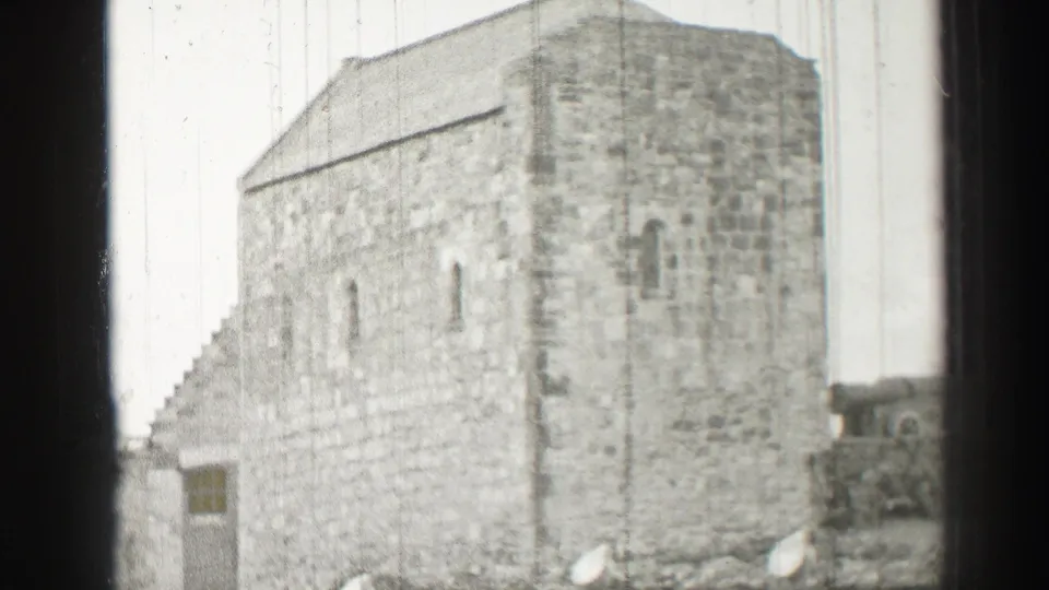 A broad exterior wall of an old stone building appears in soft black-and-white 8mm footage.
