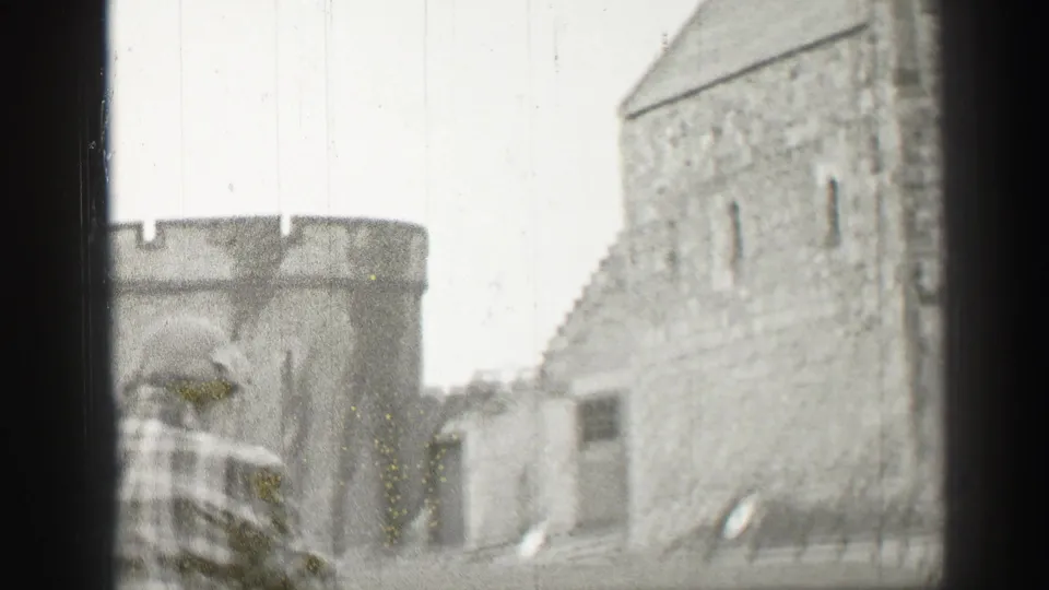 A man in traditional Scottish attire stands within Edinburgh castle grounds, with stone walls and battlements behind him.