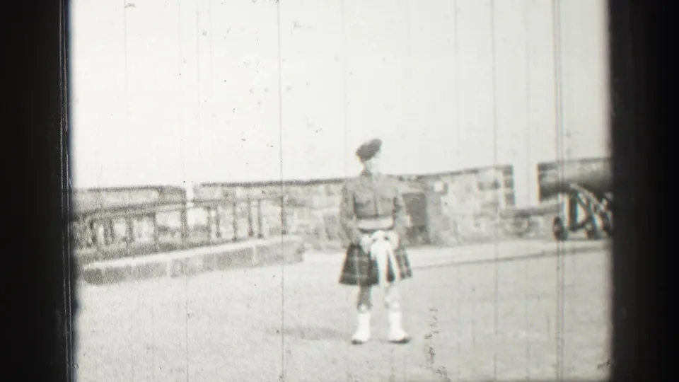 A kilted man stands on an Edinburgh castle terrace, with railings, stonework, and pale sky visible in soft 8mm focus.