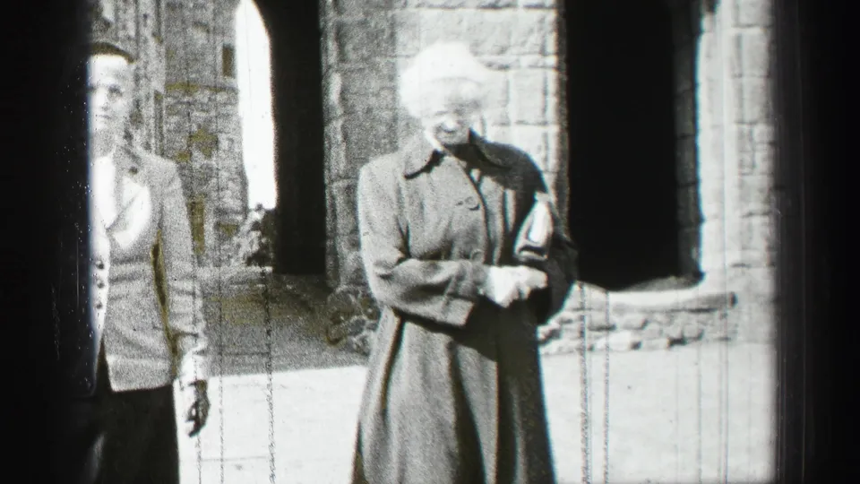 Two women in 1950s-style coats and hats walk through a stone courtyard in Scotland, with arched openings behind them.