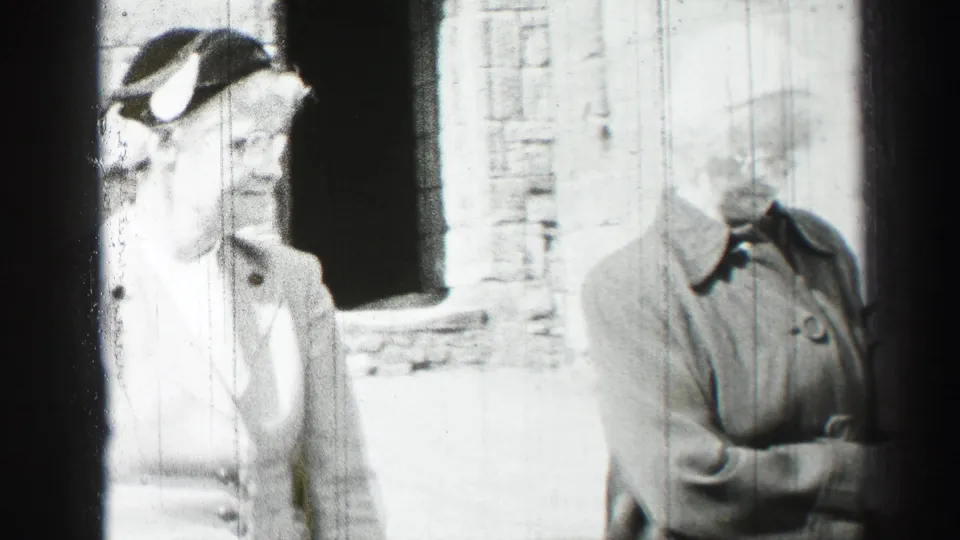 A close, overexposed home-movie view shows two women in mid-century coats and hats near a stone courtyard wall.