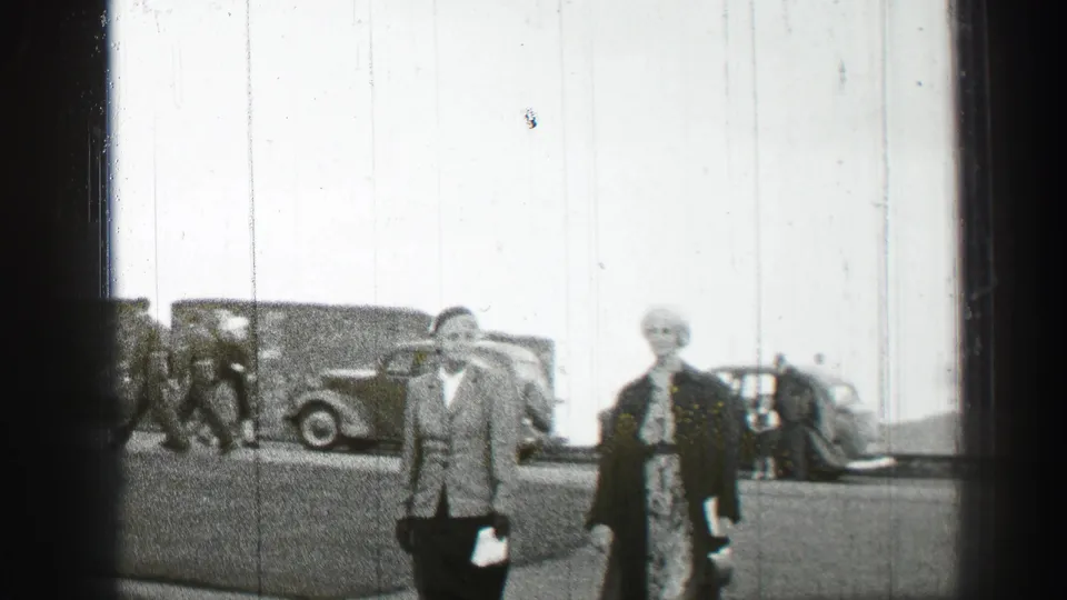 Two adults walk along a street in the supplied Scotland clip, with rounded vintage automobiles and a plain building wall behind them.
