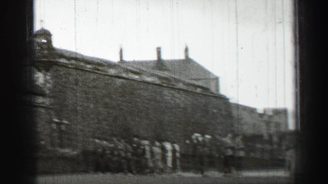 A stone wall, rooftops, and small groups of people are seen across a public street.