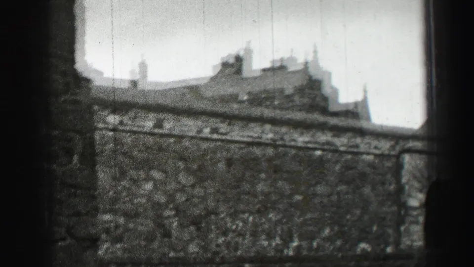 Soft black-and-white Edinburgh view of a stone wall, rooftops, chimneys, and distant pedestrians near the base.
