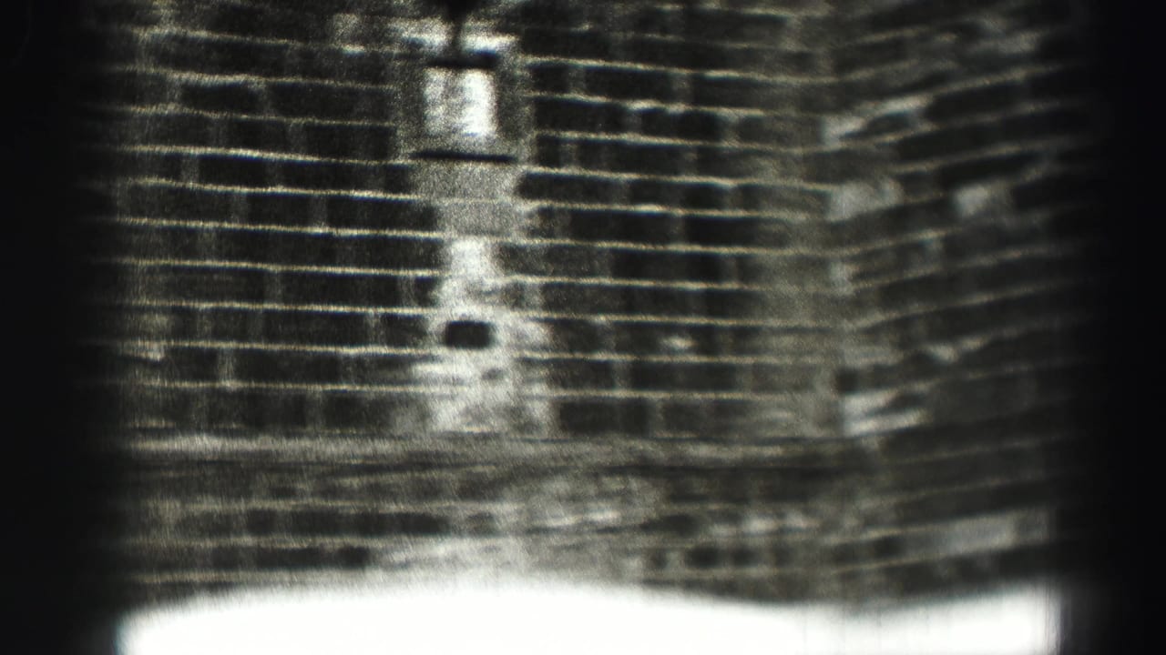 The frame shows a grainy monochrome view of a brick facade, with small wall openings and overexposed light along the lower edge.