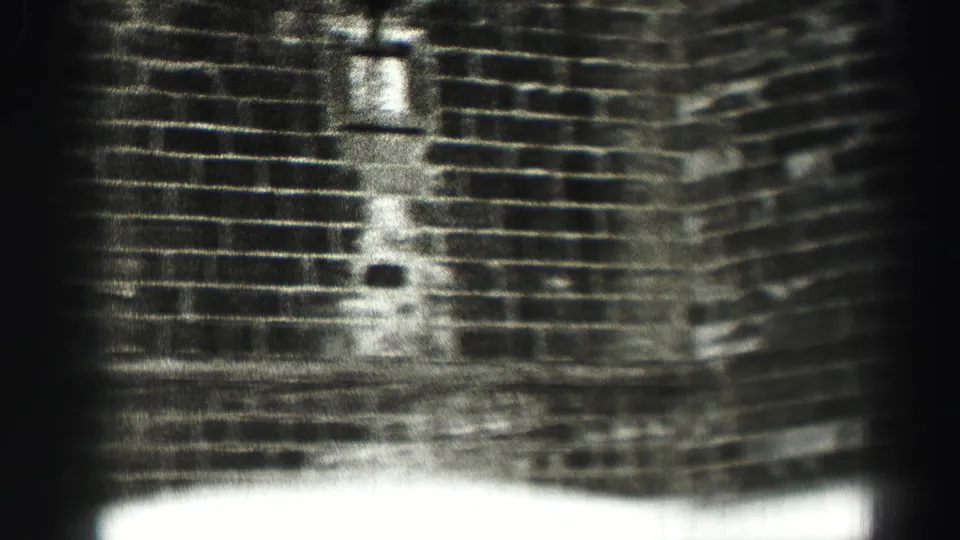The frame shows a grainy monochrome view of a brick facade, with small wall openings and overexposed light along the lower edge.