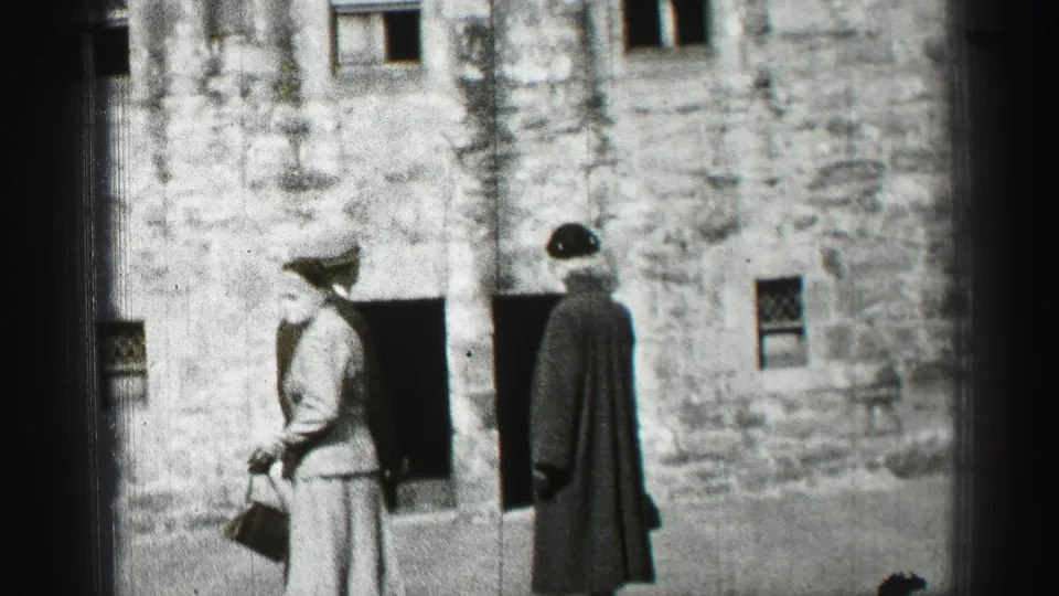 A woman in a skirt suit carrying a handbag walks past a man in a long coat outside the Edinburgh stone building.