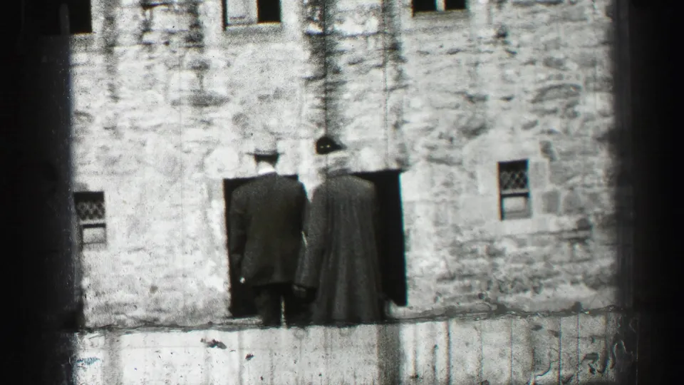Two men in hats and long coats face a weathered stone building in the 1952 Edinburgh, Scotland footage.
