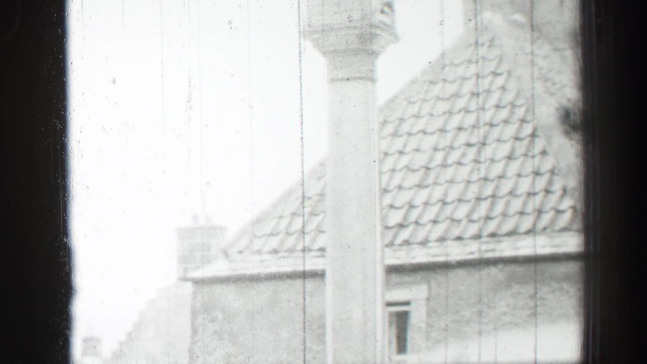 View through a window toward stone buildings with tiled roofs and a column.
