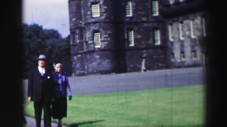 The couple continue toward the camera across the castle lawn, with rows of windows and a paved drive in the background.