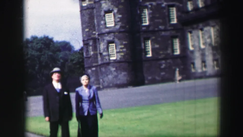 A man in a dark suit and hat and a woman in blue are framed near the camera on Edinburgh castle grounds, 1952.