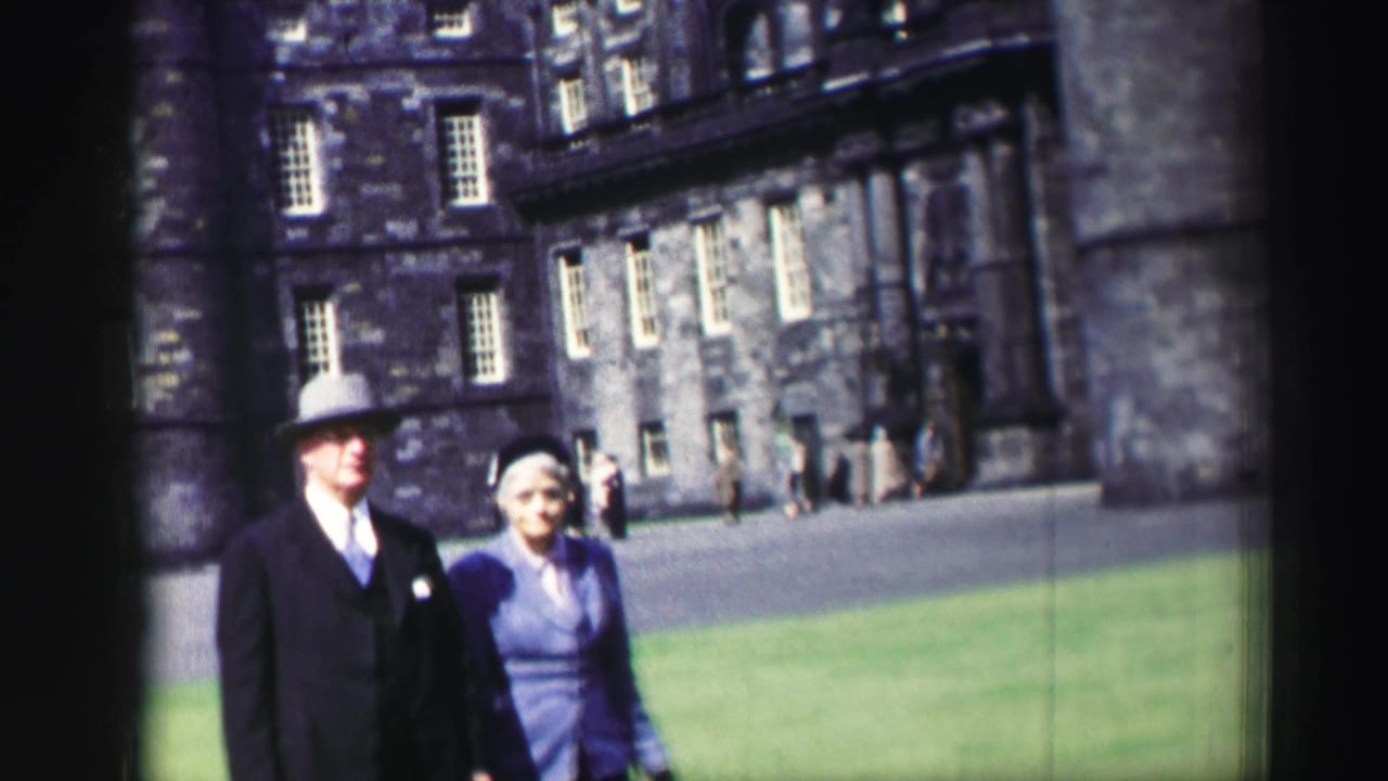 The man in a dark suit and hat walks beside a woman in purple clothing on historic castle grounds in Edinburgh.