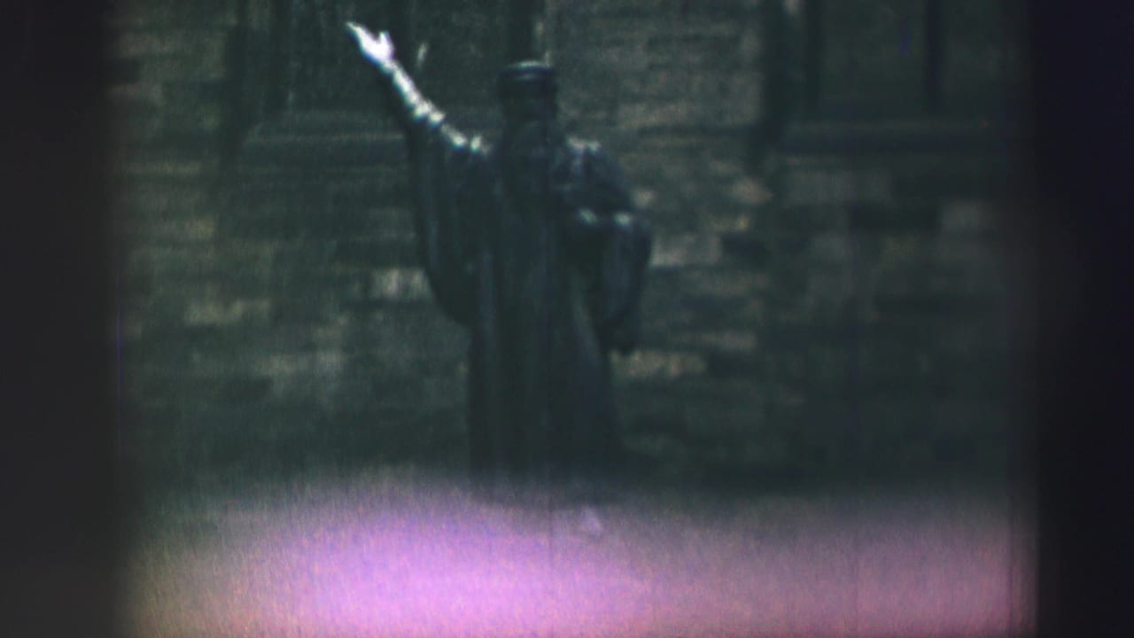 Soft 8mm frame of a dark robed statue pointing upward against a stone wall.