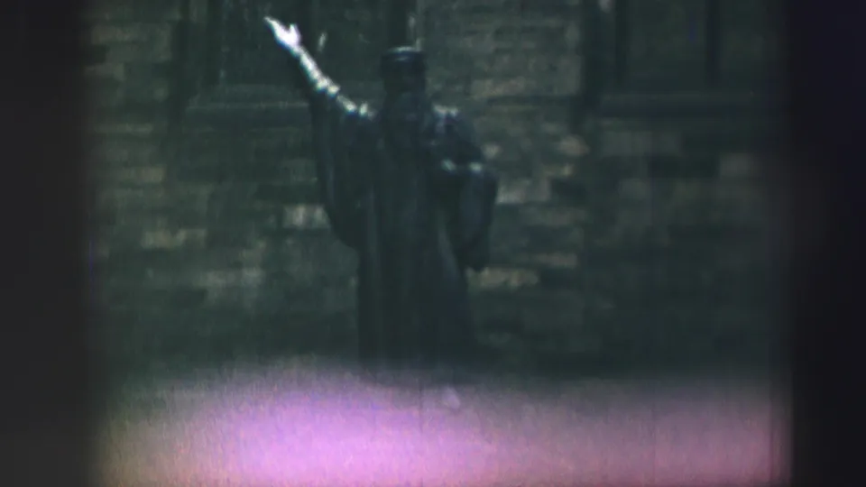 1952 Edinburgh home-movie frame showing a robed statue with one arm raised before a textured stone wall.