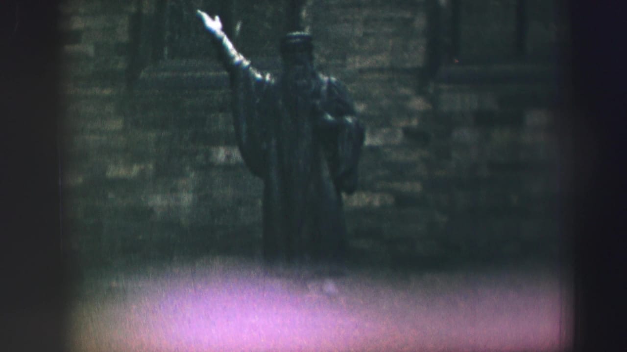 1952 Edinburgh frame holding on the raised-arm statue, with grain, dark stone background, and purple flare near the lower edge.