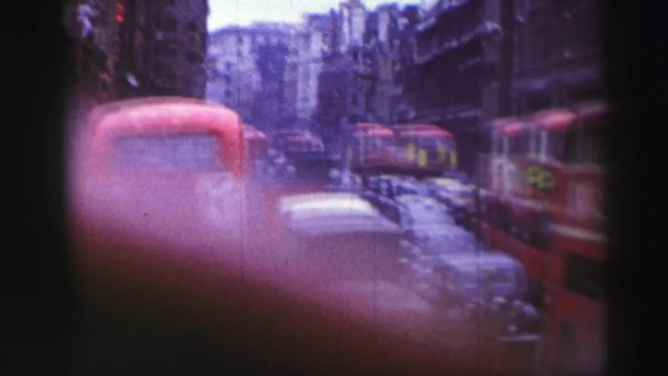 Home-movie frame of 1952 British city traffic with red double-decker buses, vintage cars, and multi-story buildings.