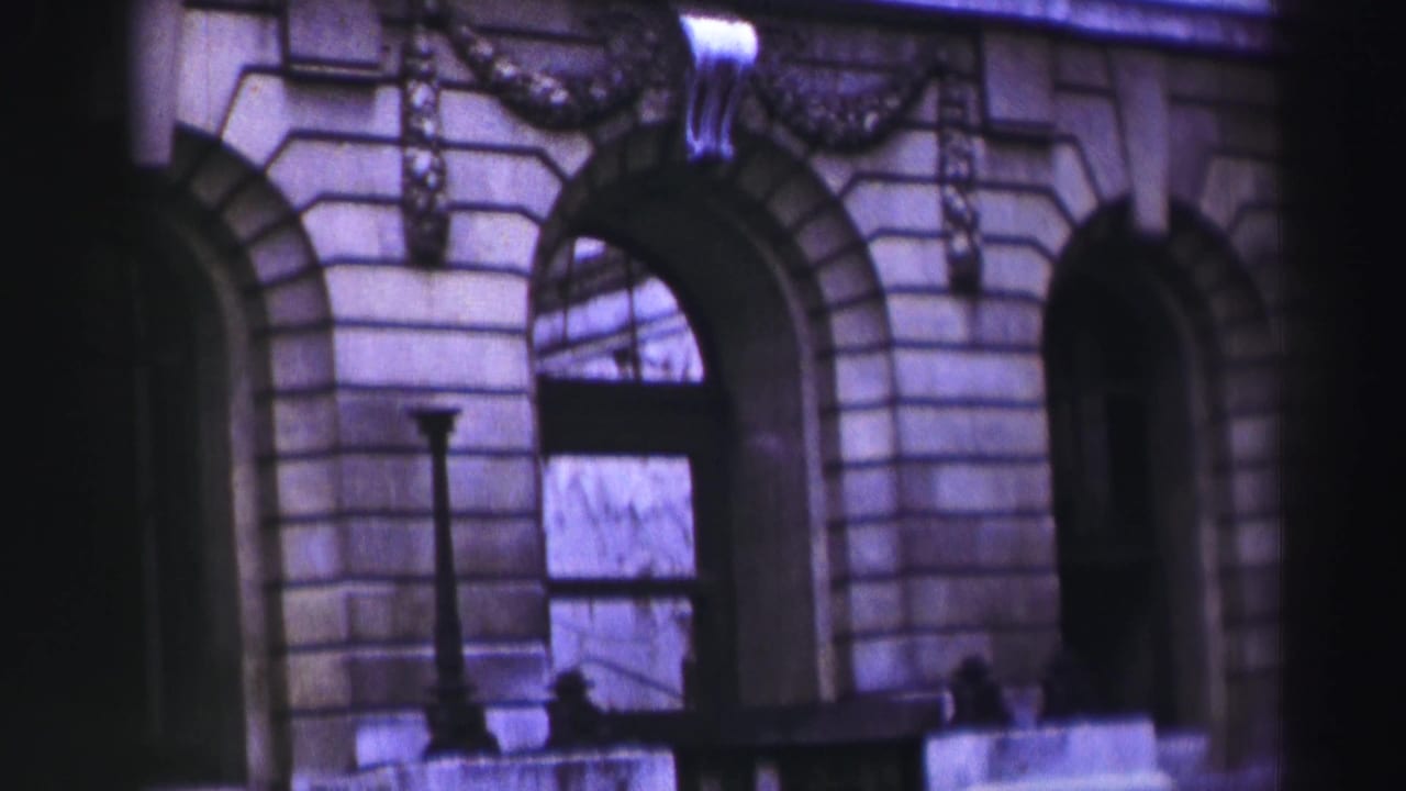 1952 8mm frame of a grand stone facade, possibly in the recorded UK travel sequence, with arches, masonry courses, and soft purple color shift.