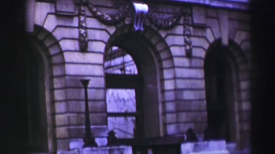 1952 8mm frame of a grand stone facade, possibly in the recorded UK travel sequence, with arches, masonry courses, and soft purple color shift.