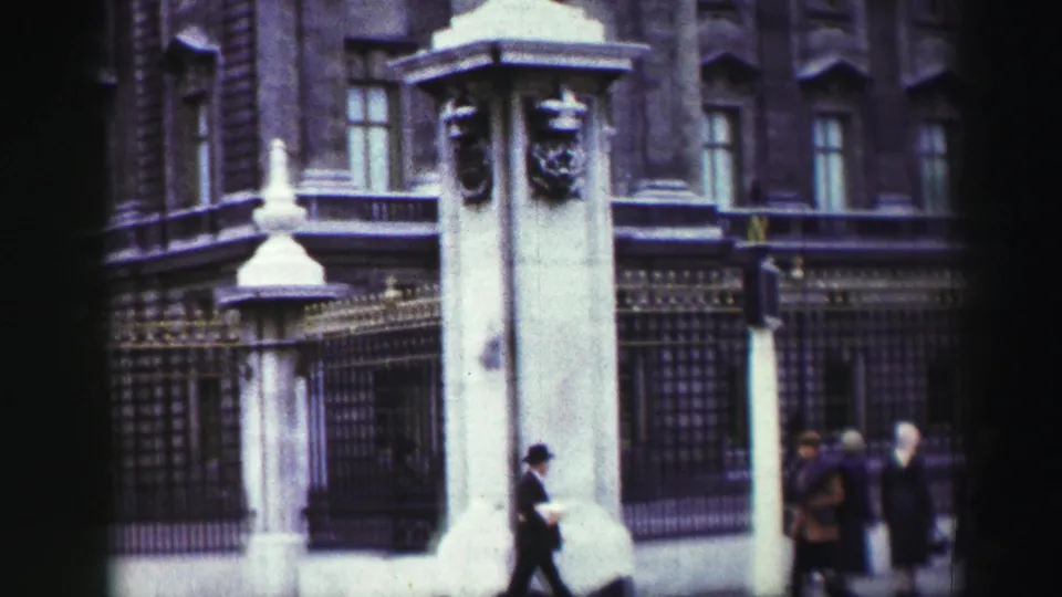 Buckingham Palace exterior appears with stone gate pillars, iron railings, tall windows, and pedestrians near the street.