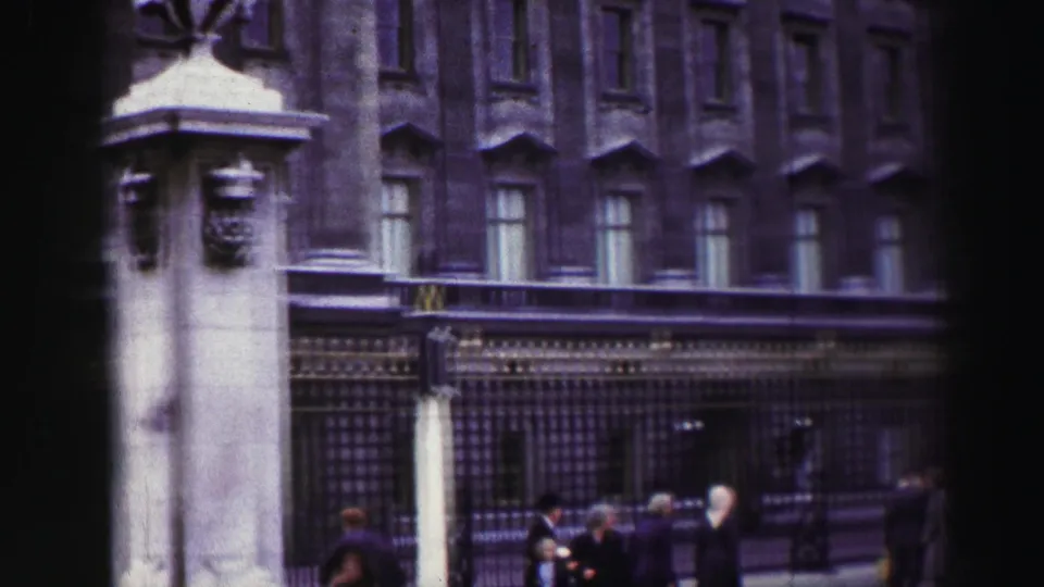 Street-level view of the palace facade, iron railings, gate pillars, and small pedestrians in 1950s home-movie footage.