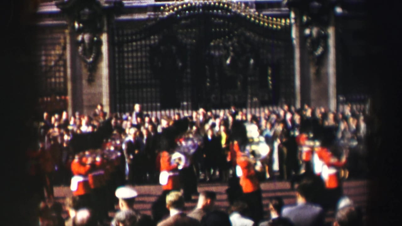 Reported 1952 Scotland frame with guards in red tunics and bearskin hats marching in front of a large crowd and ornate gate.