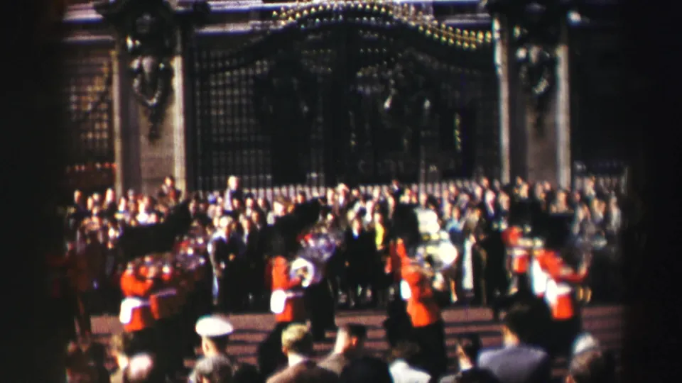 Reported 1952 Scotland frame with guards in red tunics and bearskin hats marching in front of a large crowd and ornate gate.
