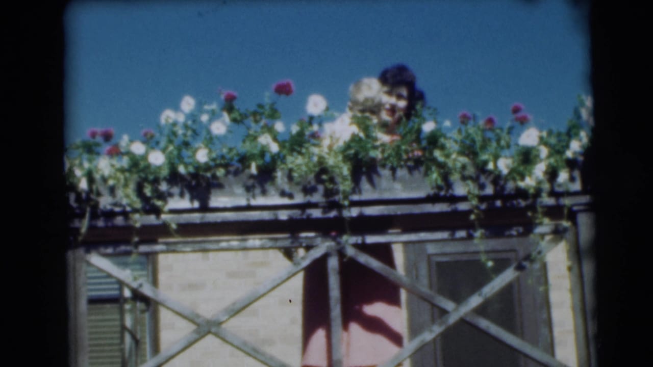 Woman holding a baby partly hidden behind flowers on a balcony under a blue sky.