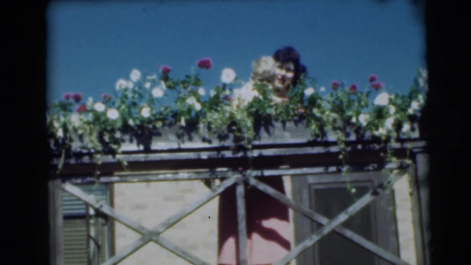 In the 1954 Pennington home movie, a woman holding a baby appears partly obscured behind a balcony flower box.