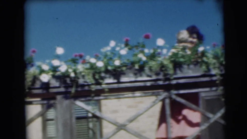1954 Pennington frame with a woman holding a baby behind bright balcony flowers and an X-braced railing.