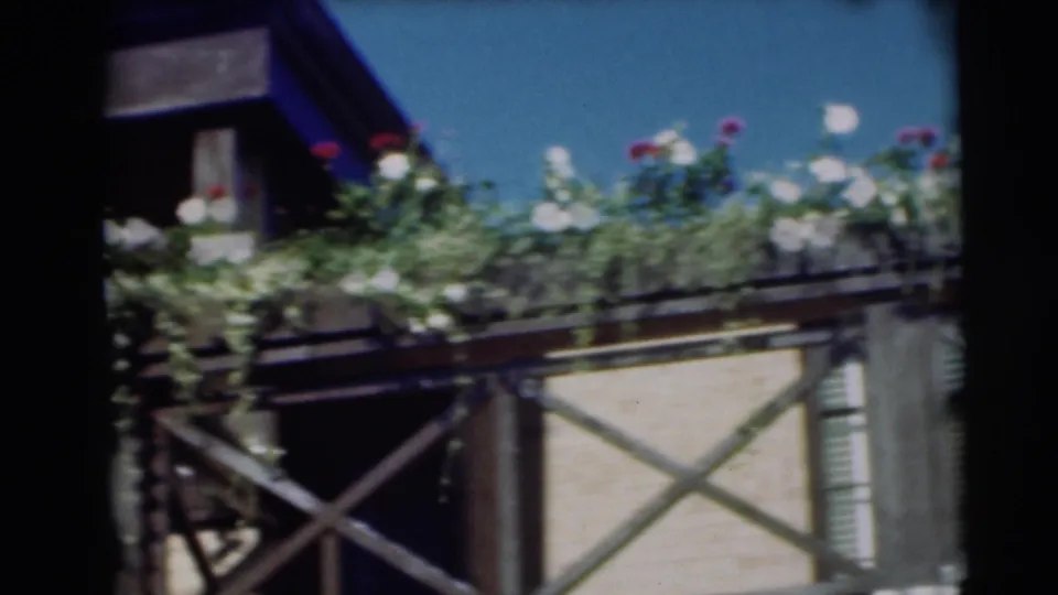 1954 Pennington home-movie frame showing balcony flowers, a pale facade, X-braced railing, and adjacent window detail.