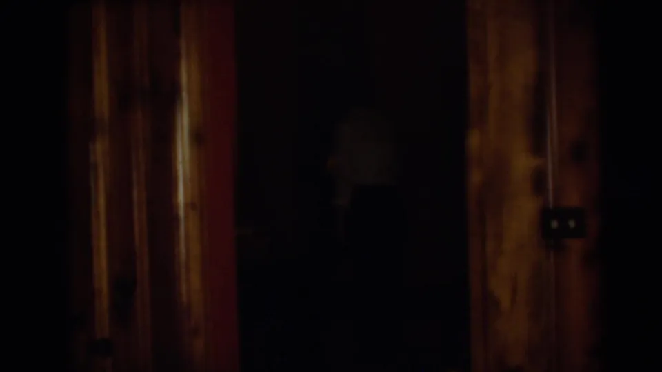 1954 Pennington home-movie frame showing a dim private interior with wood doorframes and a barely visible woman in shadow.
