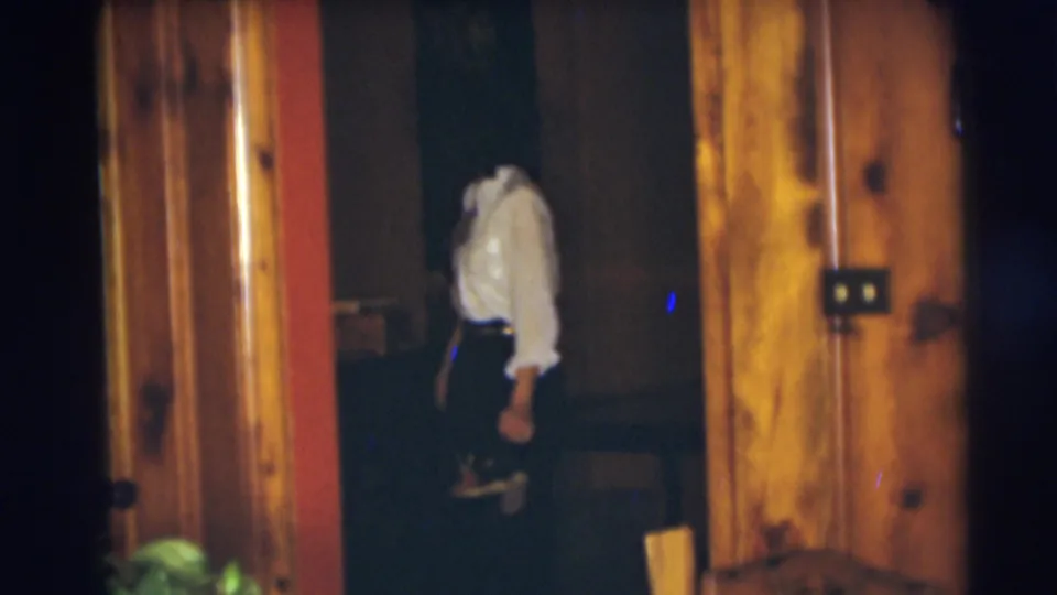 1954 Pennington frame of a woman carrying shoes through a dim private interior with wood paneling and warm household light.