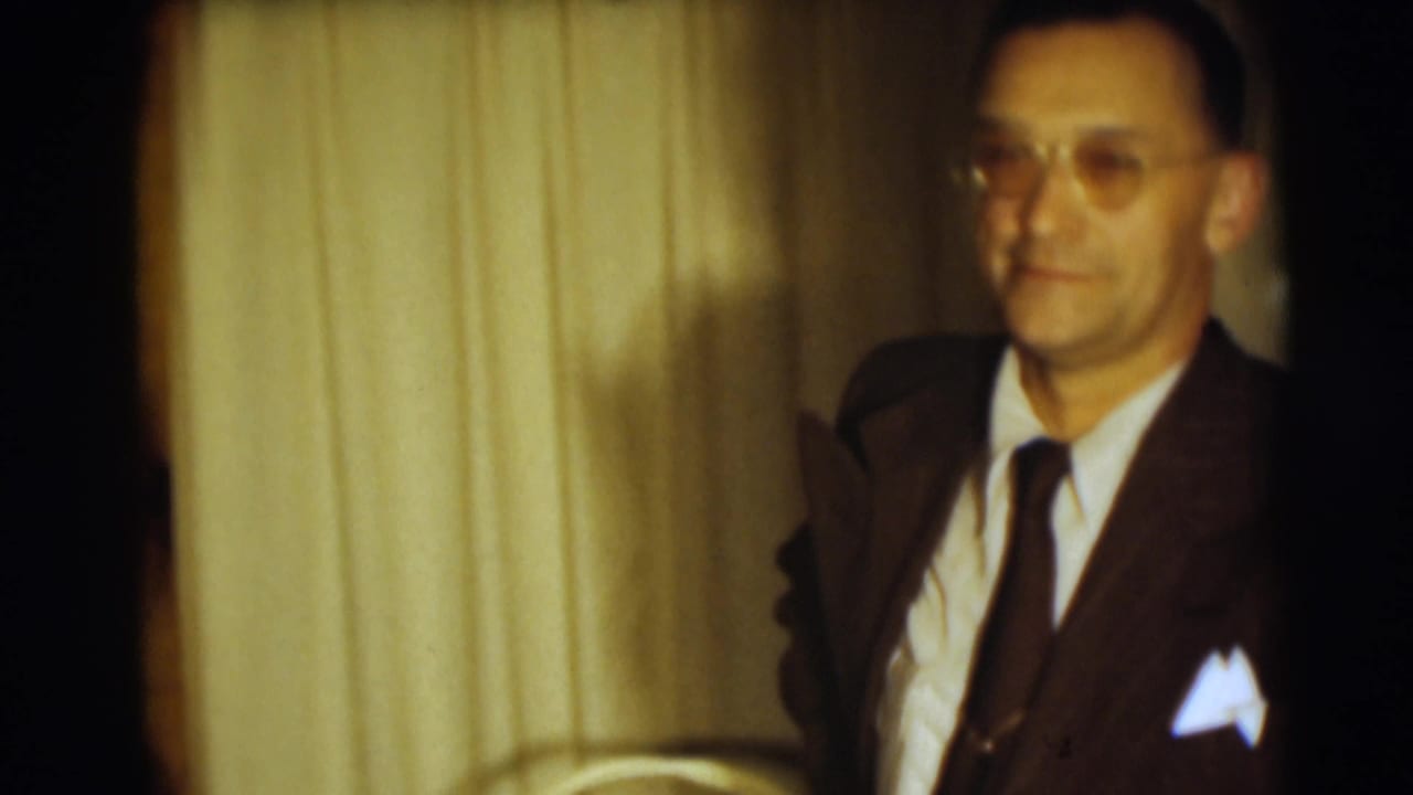 Man in a dark suit and glasses stands before curtains in a softly lit interior.
