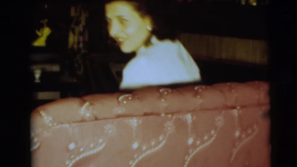 The seated woman turns toward the camera with a slight smile in the dim 1954 home interior.
