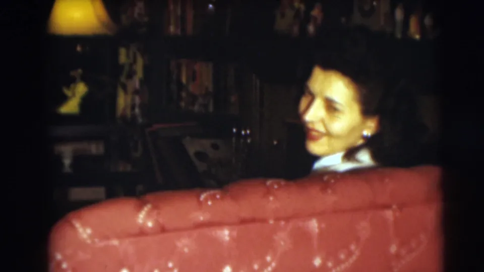 The seated woman smiles back toward the camera over a red tufted sofa in the Pennington home-movie scene.