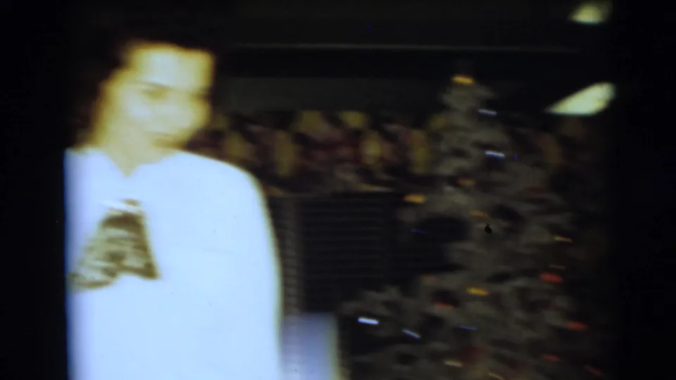 A blurred 1954 home-movie frame shows a woman in a white blouse near a decorated Christmas tree in a private interior.