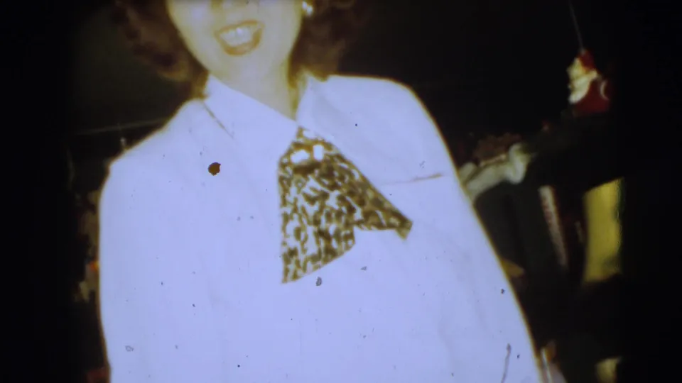 Close 1954 home-movie view of a woman in a white blouse and patterned neck scarf, with a dim holiday interior behind her.