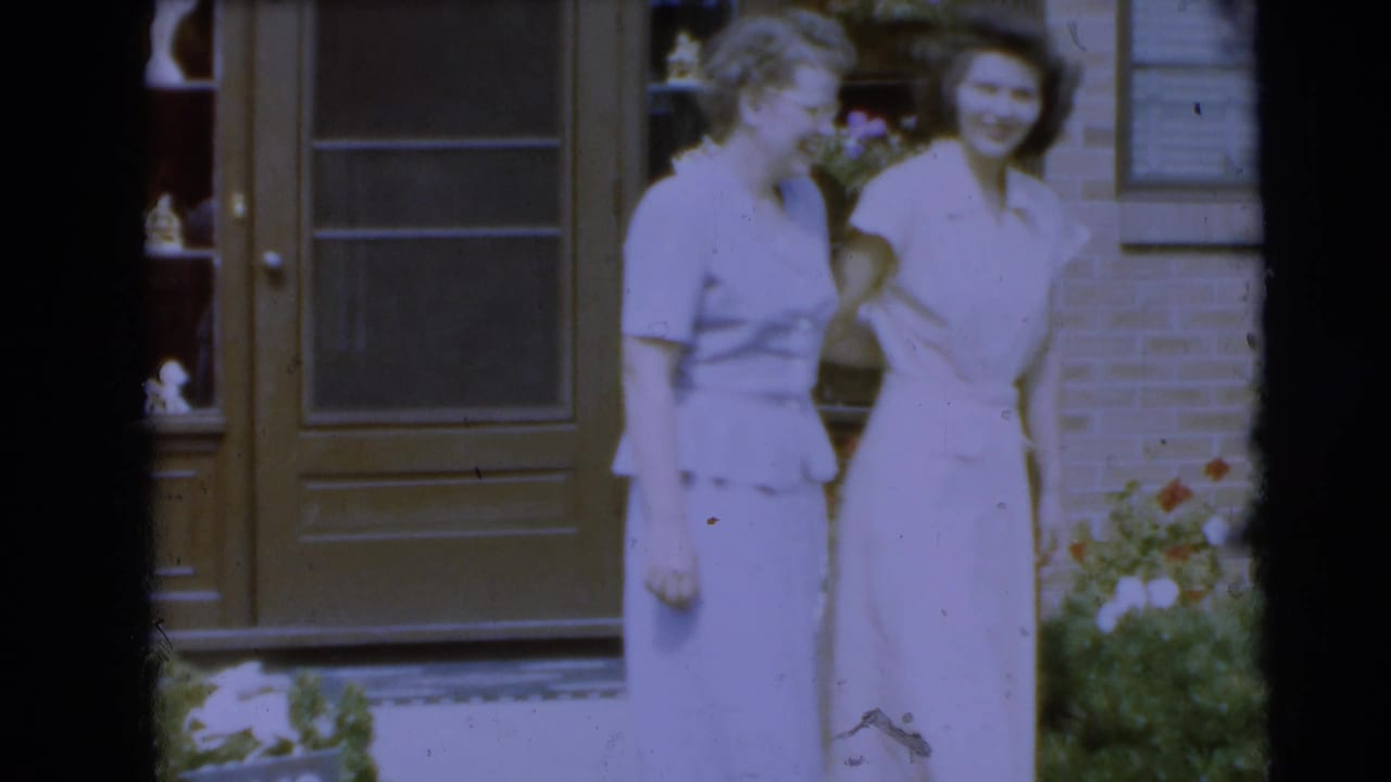 Two young women in light dresses stand outside a brick house near flowers.