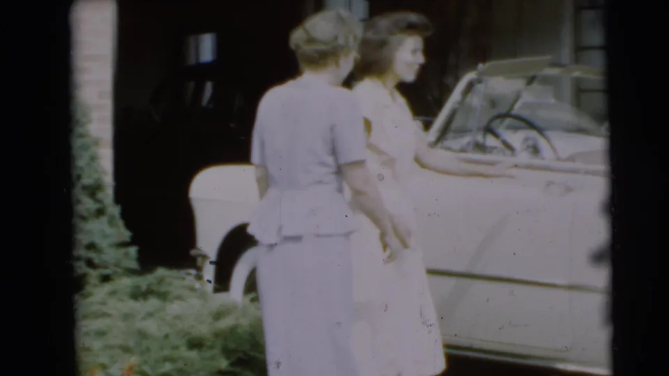 Two women in light dresses stand beside a pale convertible outside the Pennington residence in 1954.