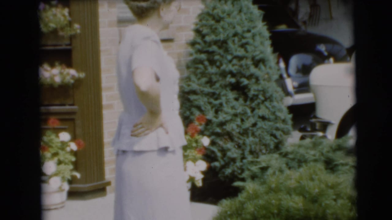 Woman in a pale dress stands near a brick house, shrubs, flowers, and a white convertible.