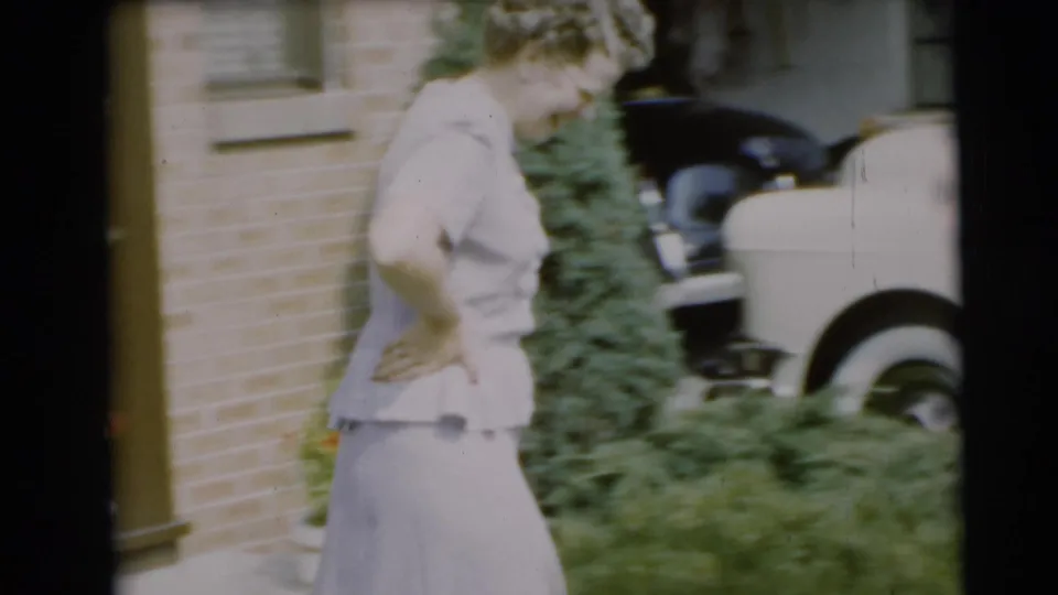 The woman smiles and moves through a Pennington yard toward the white convertible, with brickwork and greenery behind her.