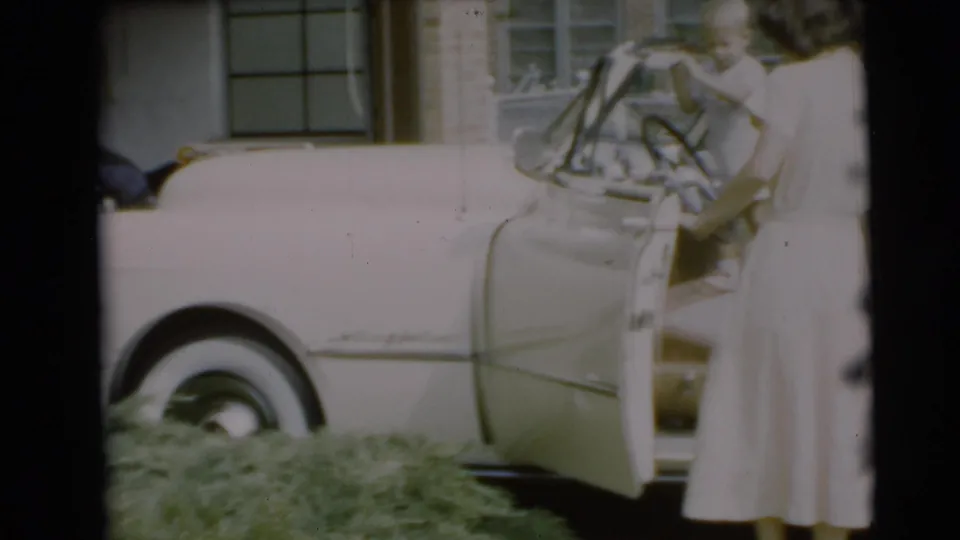 The woman stands beside the white convertible with its side open, framed by the lawn, house windows, and soft 8mm exposure.