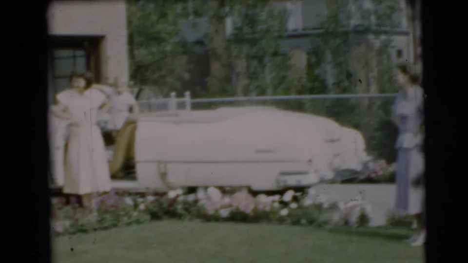 A woman stands near the white convertible in the landscaped yard, with another adult at the edge of the frame and the house behind them.