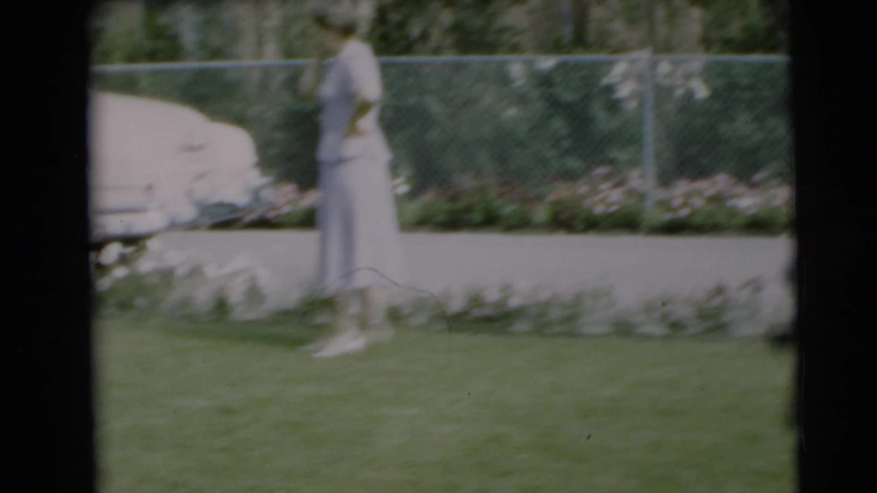 In a 1954 Pennington, New Jersey yard, a woman in a pale dress stands near a landscaped flower border and chain-link fence.