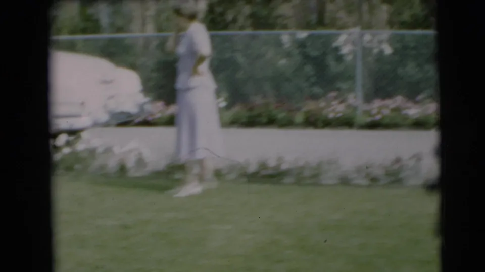 In a 1954 Pennington, New Jersey yard, a woman in a pale dress stands near a landscaped flower border and chain-link fence.